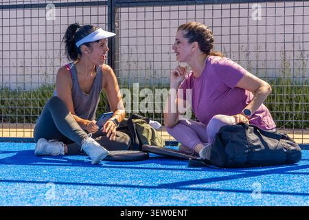 Diverse amiche che siedono e parlano sul campo blu vicino a una recinzione metallica con smartphone e pagaie Foto Stock