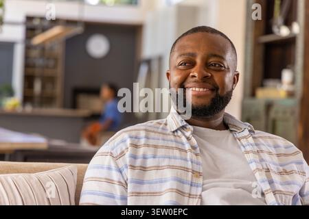 Uomo adulto afroamericano seduto sul divano in soggiorno accanto al bancone della cucina sorridente, spazio fotocopie Foto Stock