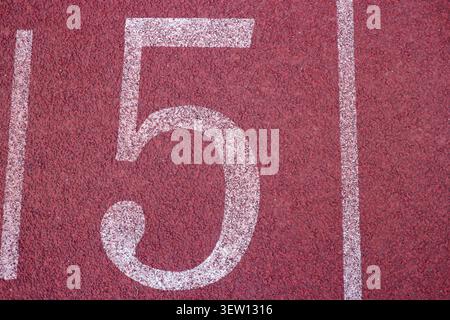 Tiro piatto ad angolo alto di un grande numero 5 bianco dipinto su una pista da corsa in gomma rossa standard in uno stadio di atletica all'aperto. Foto Stock