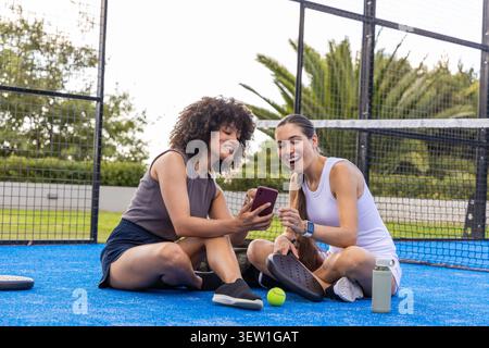 Diverse amiche di abbigliamento sportivo sedute sul campo da paddle blu che condividono lo smartphone con le pale Foto Stock
