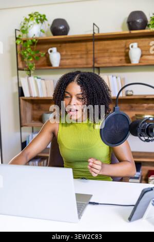 Donna afroamericana che registra in studio domestico alla scrivania con la parte superiore verde utilizzando il microfono e il computer portatile Foto Stock