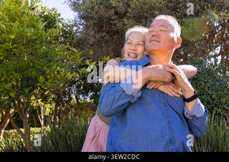 Coppia asiatica senior che abbraccia nel giardino illuminato dal sole, donna con gonna rosa, uomo con camicia in denim Foto Stock