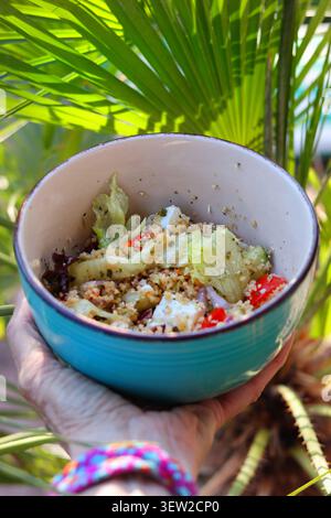 Insalata mediterranea di Couscous fresca con formaggio Feta in ciotola, all'aperto, tra la vegetazione tropicale Foto Stock