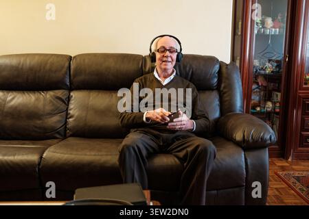 Uomo anziano che ascolta la musica con le cuffie seduto su un divano in pelle marrone a casa Foto Stock