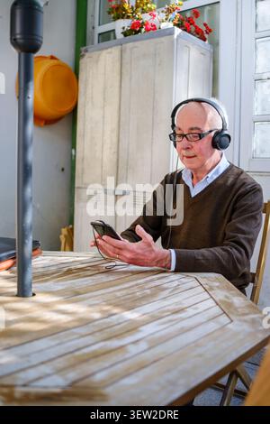 Uomo anziano seduto a un tavolo di legno, ascoltando musica con le cuffie e lo smartphone Foto Stock