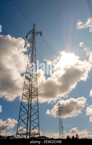 Torri di trasmissione che trasportano linee elettriche attraverso un cielo limpido con nuvole sparse e sole luminoso Foto Stock