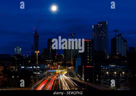 Lo skyline della città di Leeds di notte sotto la luna piena con sentieri leggeri dal traffico su una doppia carreggiata in primo piano Foto Stock
