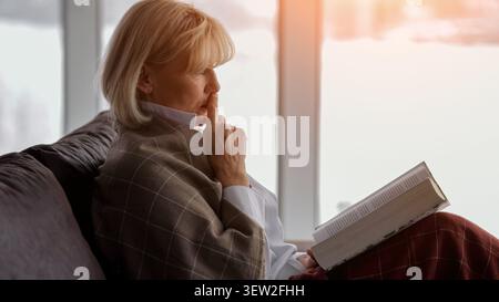 Donna anziana premurosa seduta comodamente su un divano, coperta da una coperta a quadri, che contempla mentre legge un libro alla luce della finestra, godendo di un Foto Stock