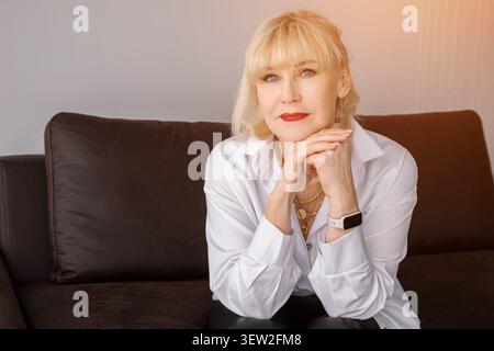 Donna anziana con capelli biondi e rossetto rosso seduta su un divano scuro, che esprime fiducia ed eleganza in un ritratto che promuove la bellezza matura Foto Stock