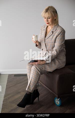 Elegante donna d'affari matura che si rilassa a casa o aspetta in una sala d'ufficio, legge una rivista e sorseggia una tazza di caffè, mostrando un Foto Stock
