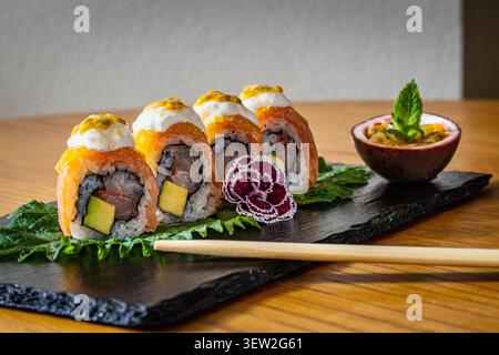 Panini di sushi con salmone, avocado e riso, serviti su una tavola di ardesia con foglie guarnite, fiori decorativi e un lato di frutto della passione. Foto Stock