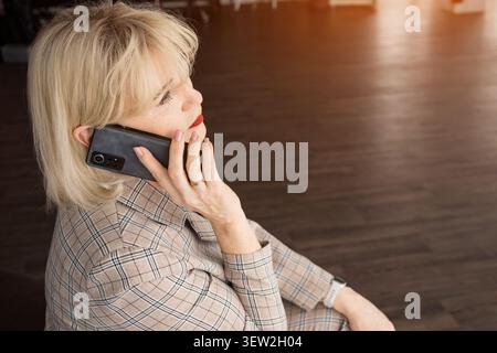 Una donna d'affari bionda matura con una tuta a quadri effettua una telefonata mirata in ufficio, illustrando comunicazioni professionali, lavoro remoto e. Foto Stock