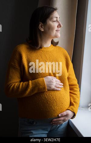 Donna incinta alla luce calda del sole vicino a una finestra che tiene la pancia Foto Stock