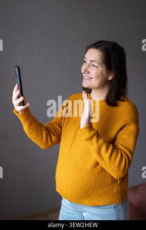 Donna incinta che tiene in mano uno smartphone e parla al telefono al chiuso Foto Stock