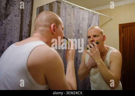 Uomo che cura il viso con un rasoio elettrico, guardando nello specchio del bagno Foto Stock