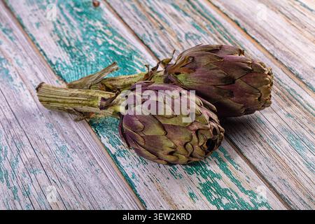Due carciofi verdi viola poggiano su legno indurito con vernice verde acqua. La luce naturale accentua il contrasto cromatico. Composizione rustica ideale Foto Stock