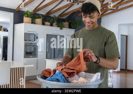 Adulto afroamericano in piedi e con abiti pieghevoli tenendo in mano il cestino della biancheria in cucina Foto Stock