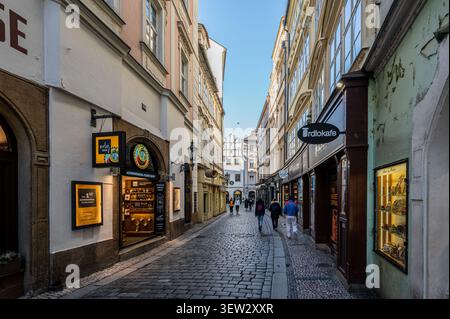 Pedoni camminano lungo una stretta strada acciottolata fiancheggiata da edifici storici e negozi locali durante il giorno a Praga, Repubblica Ceca. Foto Stock