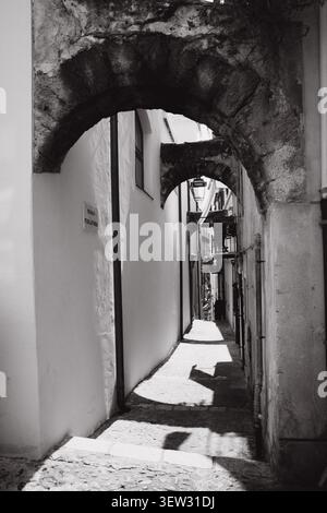 Arco in pietra bianca e nera che conduce in uno stretto vicolo illuminato dal sole con luci e ombre drammatiche su pareti bianche, passaggio storico della città vecchia in Sicilia, i Foto Stock