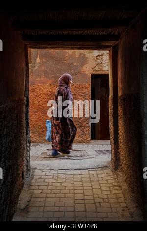 Marrakech, Marocco - 3 dicembre 2017: Una donna che indossa un hijab viola e una djellaba a motivi geometrici cammina attraverso un vicolo stretto e ombreggiato con un ruvido, t Foto Stock