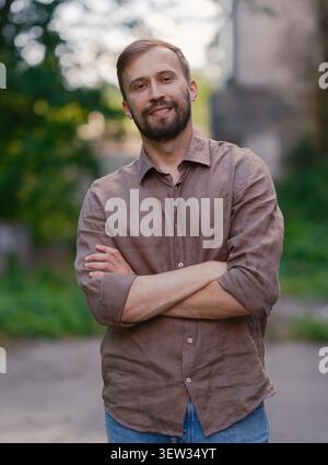 Ritratto di un uomo caucasico felice e attraente in una camicia di lino marrone casual che posa all'aperto con le braccia incrociate. Sfondo urbano sfocato con vecchio edificio, Foto Stock