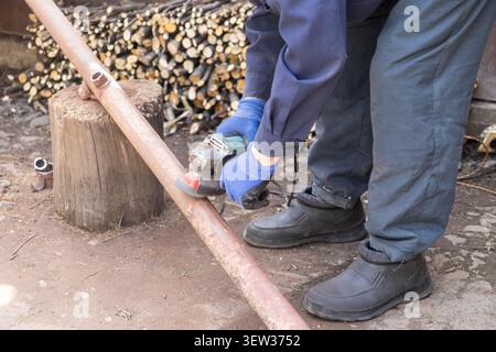 Un tuttofare che indossa guanti protettivi blu aziona un utensile elettrico per rifinire un palo in acciaio corroso, mostrando la manutenzione dettagliata della lavorazione dei metalli in una b Foto Stock