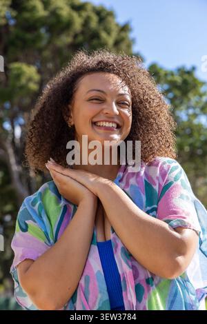 Donna afro-americana sorridente e in piedi alla luce del sole del parco, indossando una camicia colorata con motivi geometrici Foto Stock
