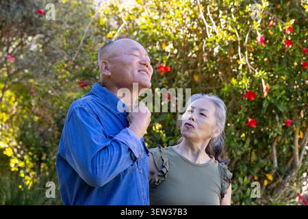 Coppia asiatica senior che posa in giardino, fiori rossi, uomo denim respira, donna top di oliva che lo tocca Foto Stock