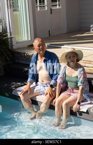 Coppia asiatica seduta a bordo piscina con piedi in acqua, con cappello di paglia, asciugamano, smartwatch Foto Stock