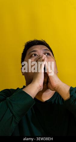 Un uomo asiatico con la barba che tiene le mani premute insieme davanti al viso con un gesto premuroso o orante contro un giallo brillante Foto Stock