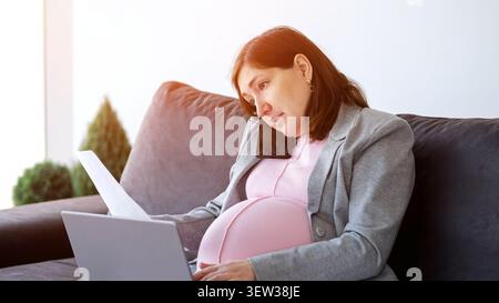 Donna incinta che lavora da casa in multitasking con telefono e laptop Foto Stock