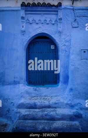 Una vivace parete dipinta di blu con una porta ad arco in legno, numerata 33, si erge sopra una serie di gradini blu usurati. La scena cattura il distintivo architec Foto Stock