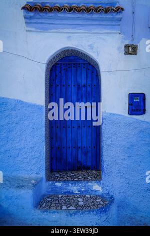 Chefchaouen, Marocco - 7 dicembre 2017: Una porta ad arco in legno blu intenso e vibrante con borchie nere è adagiata su un muro dipinto in varie tonalità di blu Foto Stock