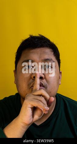 Un uomo adulto calmo che indossa una camicia verde scuro tiene il dito indice sulle labbra con un gesto tranquillo e scintillante su uno sfondo giallo chiaro e solido. Foto Stock