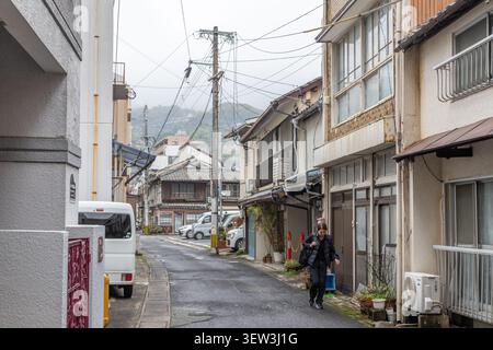 Stretta via residenziale a Nagasaki, Giappone, fiancheggiata da edifici più antichi, veicoli parcheggiati e cavi sospesi. Foto Stock