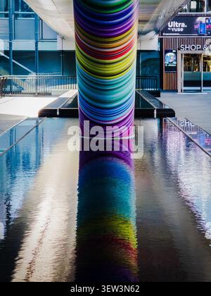 Una colonna a strisce arcobaleno dell'opera d'arte di Adam Nathaniel Furman "Click Your Heels Together Three Times" riflessa in una piscina d'acqua a Canary Wharf, Londra Foto Stock