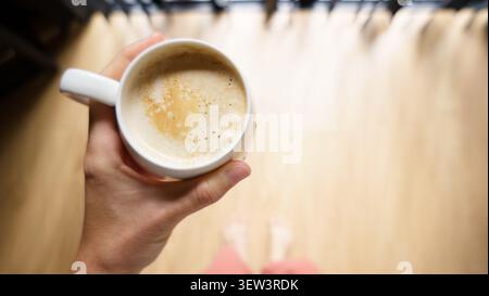 Persona che inizia la giornata tenendo il caffè, stando a piedi nudi nella calda luce del mattino a casa Foto Stock