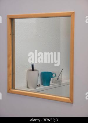 Vista ravvicinata dello specchio del bagno con cornice in legno che riflette lo scaffale compatto con porta spazzolino bianco, spazzolino nero, tazza in ceramica color tè, vetro trasparente, sp Foto Stock