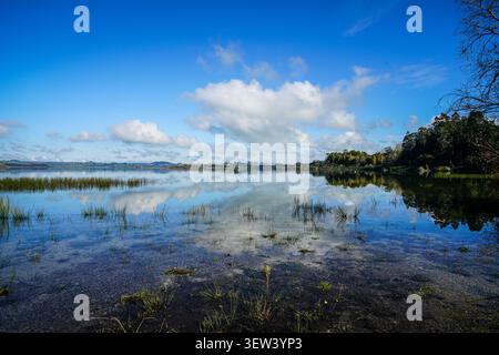 Una fotografia di paesaggio grandangolare cattura un lago tranquillo o un grande specchio d'acqua sotto un cielo blu luminoso con nuvole bianche sparse, situate sul Foto Stock