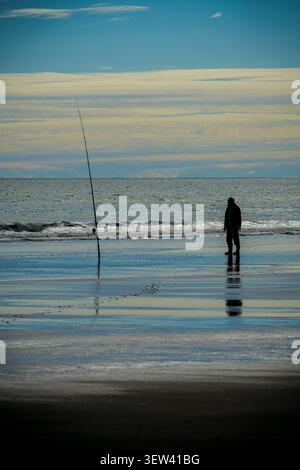 Un pescatore solitario si staglia contro il cielo luminoso, in piedi su un'ampia spiaggia bagnata accanto a una lunga canna da pesca bloccata nella sabbia. Piccole onde rotolano Foto Stock