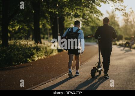 Due amici camminano in un parco al tramonto con uno scooter elettrico su un sentiero alberato Foto Stock