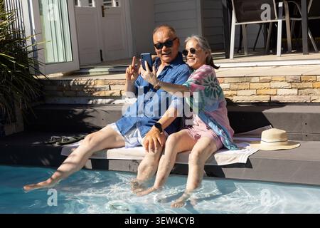 Coppia asiatica senior seduta sul ponte di legno a bordo piscina sul retro, scattando selfie con lo smartphone Foto Stock