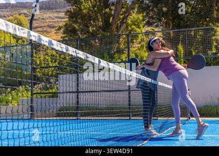 Diverse amiche che abbracciano la rete sul campo del padel con abbigliamento sportivo che tiene le pagaie e lo spazio fotocopie Foto Stock