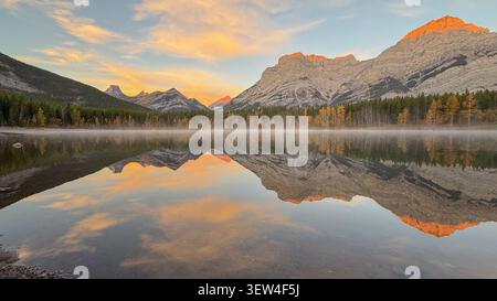 Riflesso dell'alba del Monte Lawrence grassi sulle acque calme del laghetto Wedge circondato da alberi autunnali nella regione di Kananaskis, Alberta, Canada Foto Stock