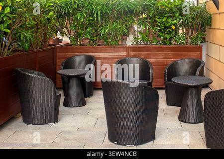 Confortevole arredamento esterno in rattan con poltrone e tavoli in vimini in un accogliente patio circondato da piante verdi in fioriere di legno. Foto Stock