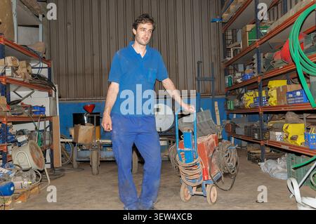 Un operaio di fabbrica maschile in uniforme blu spinge un carrello per attrezzature pesanti all'interno di un magazzino pieno di utensili, cavi e scaffali industriali. Foto Stock