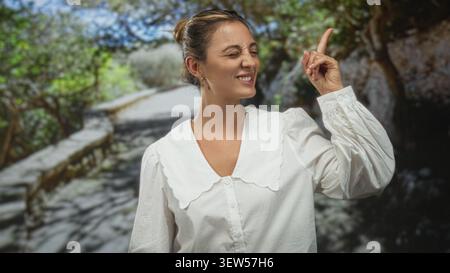 La donna punta il dito indice verso l'alto sul sentiero della foresta, gli occhi chiusi e sorridenti in camicetta bianca; momento di gioia. Foto Stock