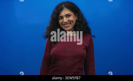 Giovane donna bruna sorridente con volto visibile e postura rilassata in studio blu, indossa un maglione rosso e guarda verso la fotocamera; gioia. Foto Stock