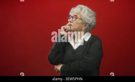 Donna anziana che indossa occhiali con la mano sul mento e le braccia piegate in uno studio rosso, camicia con colletto bianco e maglione nero; riflesso riflesso riflesso riflesso. Foto Stock
