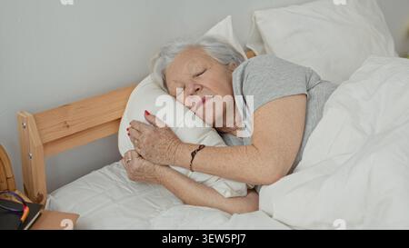 Una donna anziana abbraccia un cuscino mentre dorme su un letto in una camera da letto, distesa sul fianco con gli occhi chiusi e le mani che stringono il cuscino; comfort serenità. Foto Stock